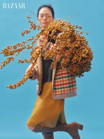 Fashion editorial shot: Woman in checkered jacket and boots poses with dried seed pods against a vibrant turquoise background.