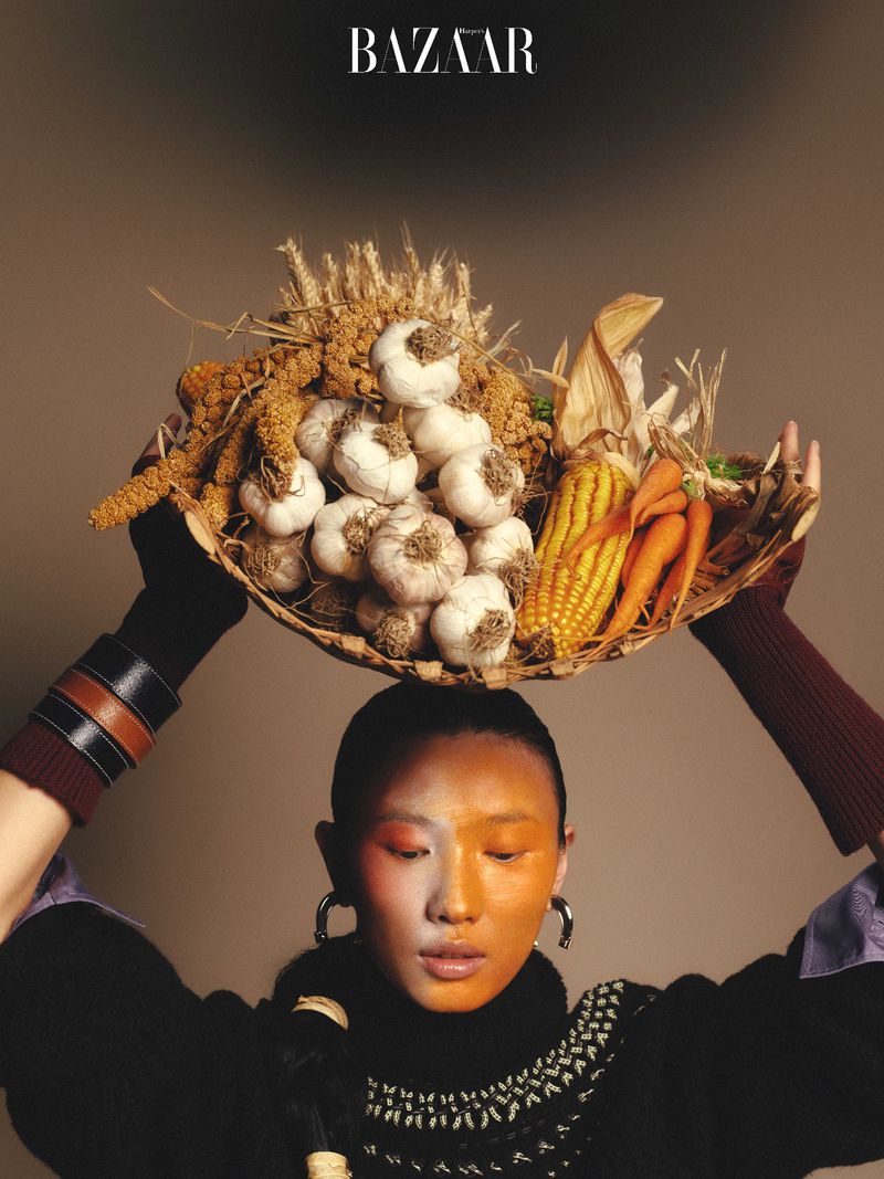 Fashion editorial shot of a woman with face paint holding a basket of garlic, corn, and wheat with a tan-colored background.
