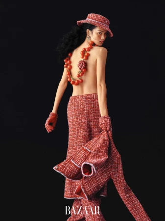 Model wearing a red tweed hat, tailored pants, and a cherry tomato back necklace posing against a black background.