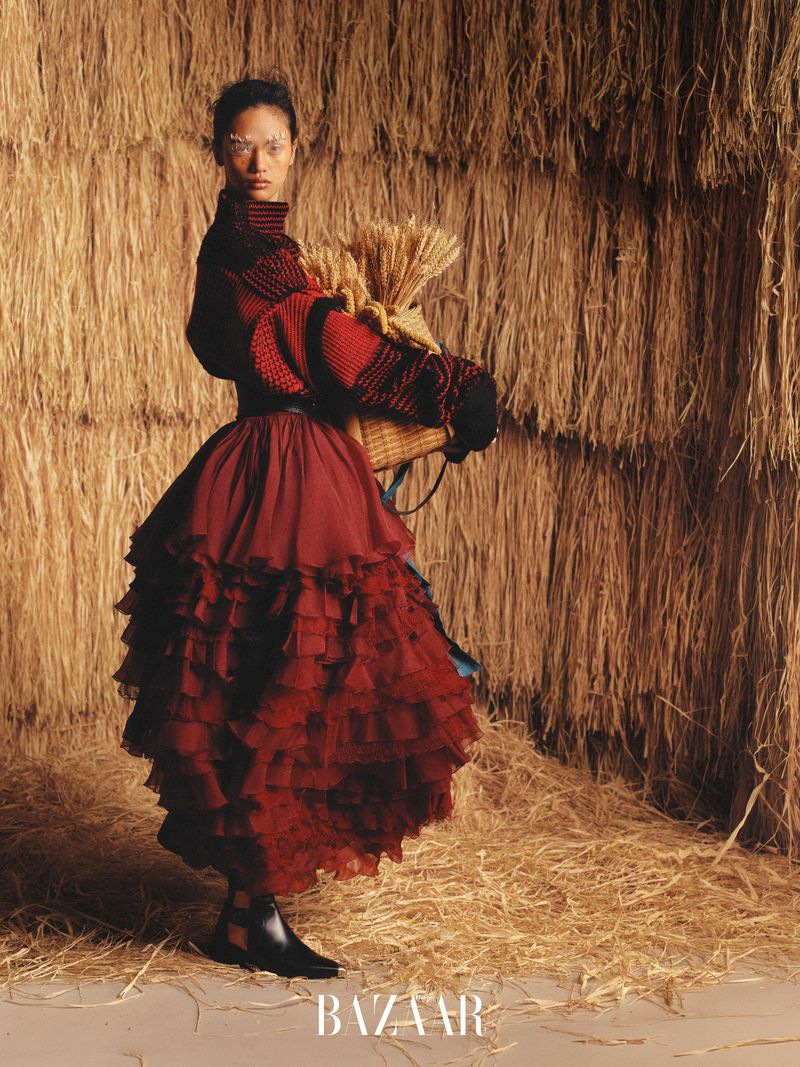 Fashion shot of a woman in a red ruffled dress carrying wheat in a woven basket against a hay bale background in a photo studio.