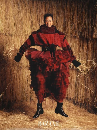 Fashion portrait of an adult woman wearing a red knit top and a ruffled skirt posing against a backdrop of bundled straw.