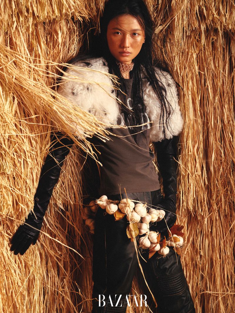 A fashion shot featuring an Asian model in black leather gloves and pants, a fur shawl, and a garlic necklace, posing against hay.