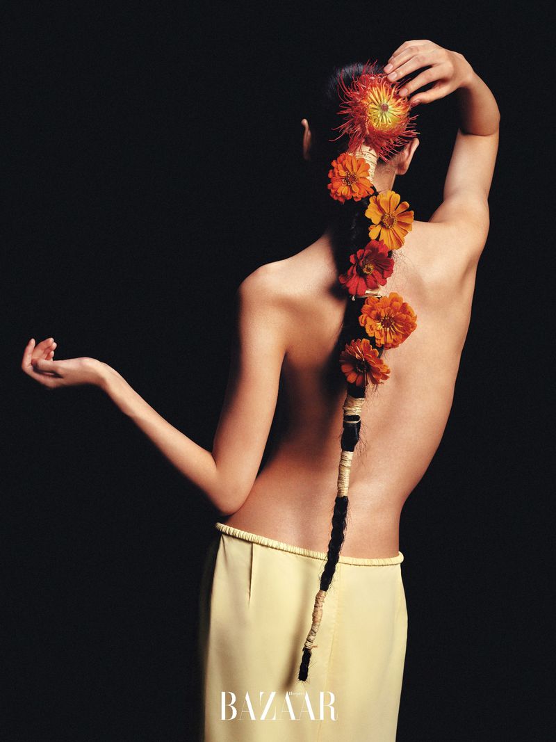 Back view of a slender woman with a long braid decorated with orange flowers, against a dark background. Fashion and beauty editorial.