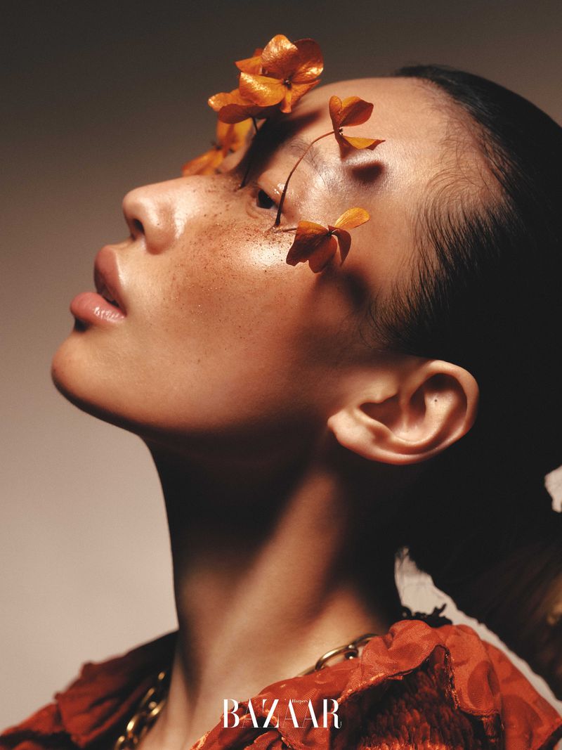 Profile shot of a woman with flowers on her face, wearing a reddish-orange top and a thick gold chain necklace, against a neutral background.