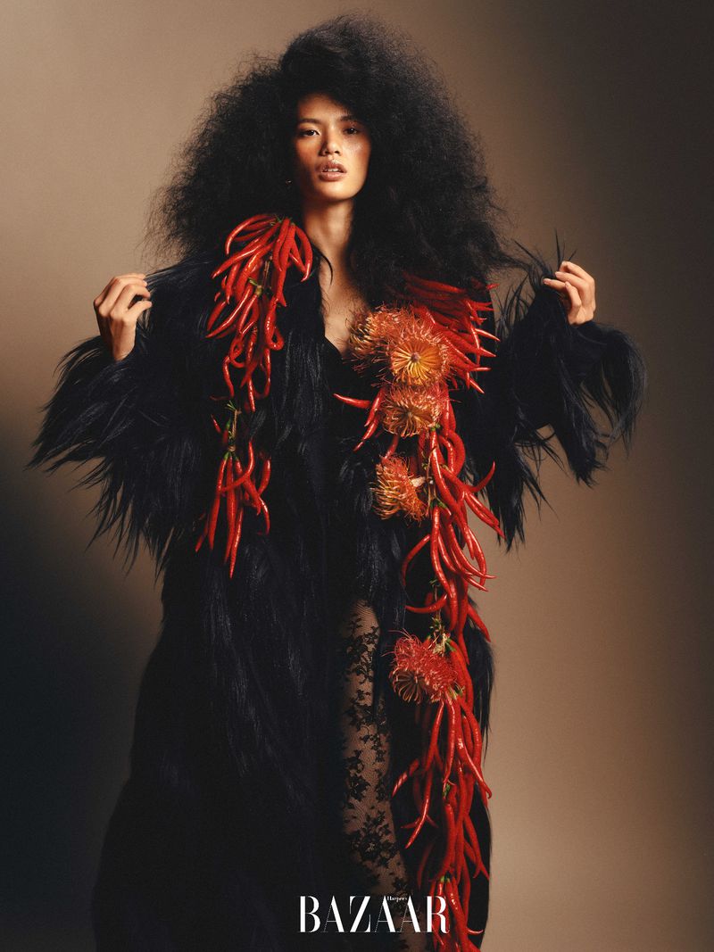 Fashion editorial portrait of an Asian woman with a large Afro, wearing a fur coat decorated with chili peppers, posing indoors.