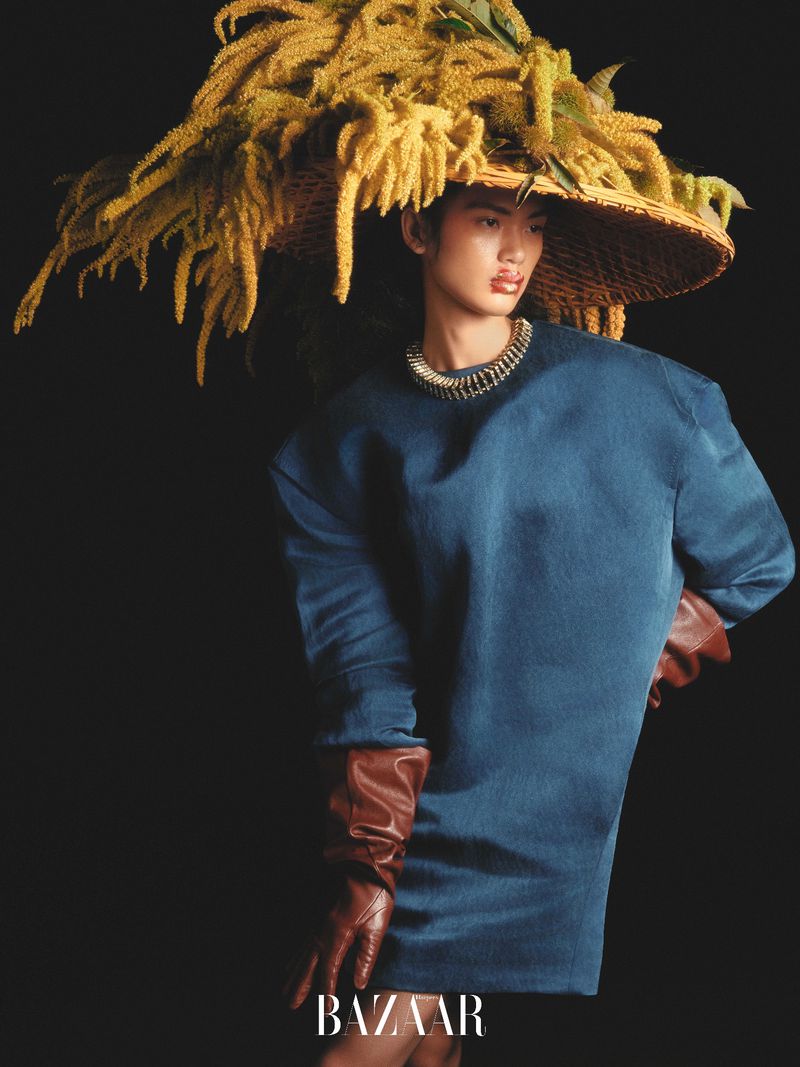Editorial fashion shot of a model in a blue dress, brown gloves, straw hat with yellow flowers, posing against a dark background.