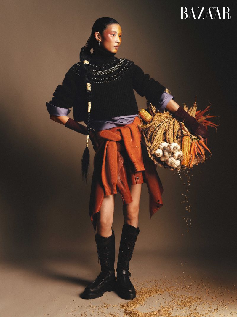 Editorial fashion shot of an Asian woman in a black sweater and tall boots, carrying a woven basket filled with corn and garlic.