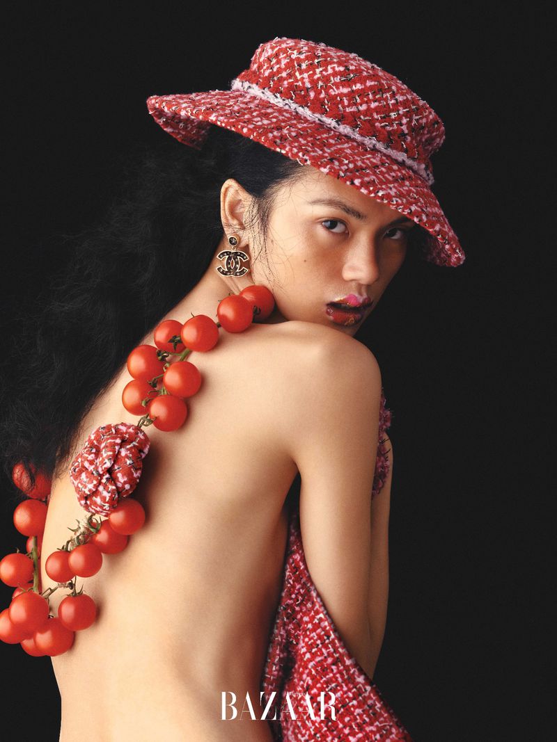 Eye-level shot of a woman with red hat and tomato necklace over her shoulder, posing against a dark background in a Bazaar fashion editorial.