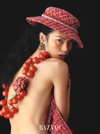 Eye-level shot of a woman with red hat and tomato necklace over her shoulder, posing against a dark background in a Bazaar fashion editorial.