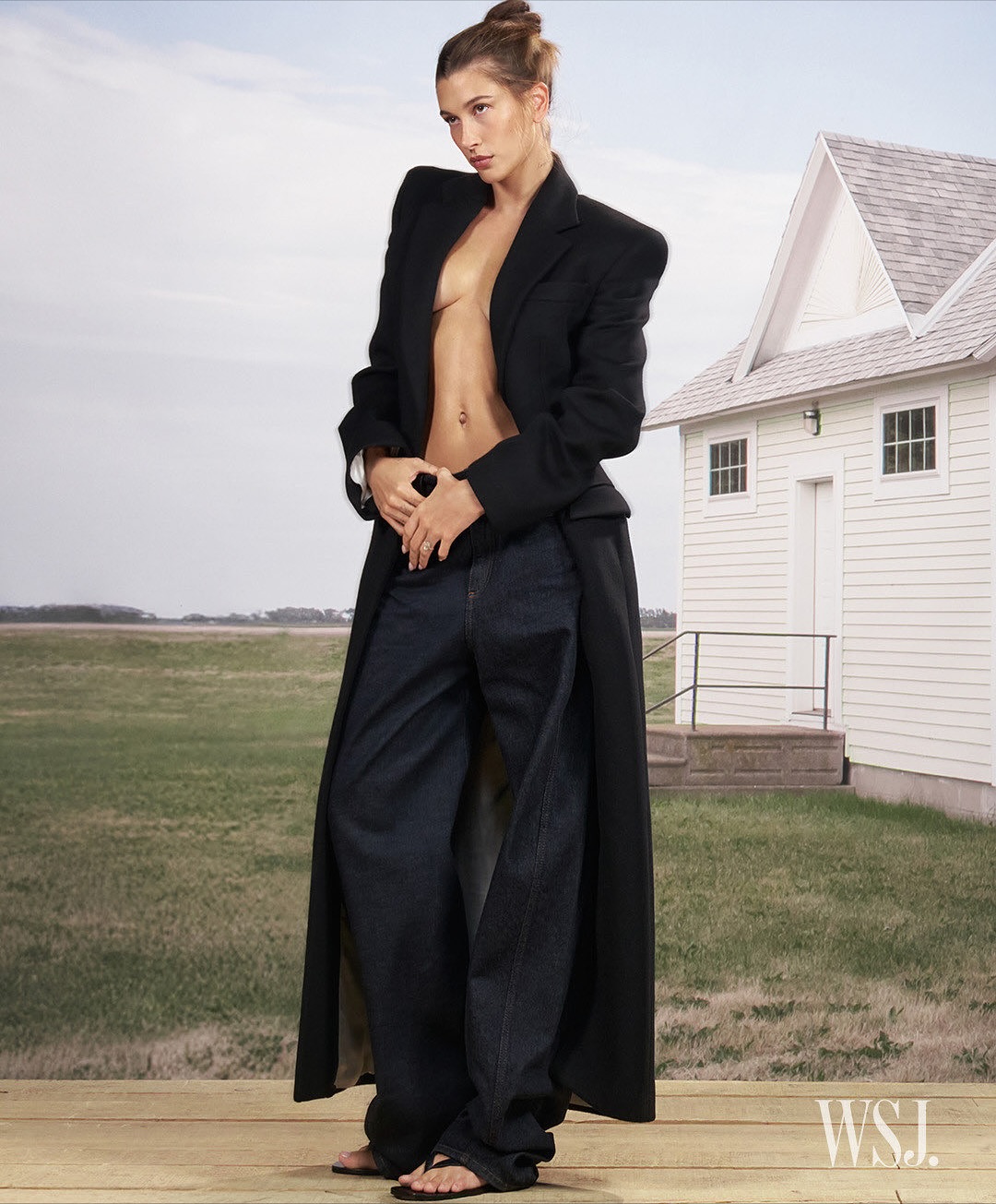 A tall, slim female model poses outdoors on a wooden deck wearing a long black coat, dark wash baggy jeans, and black sandals.