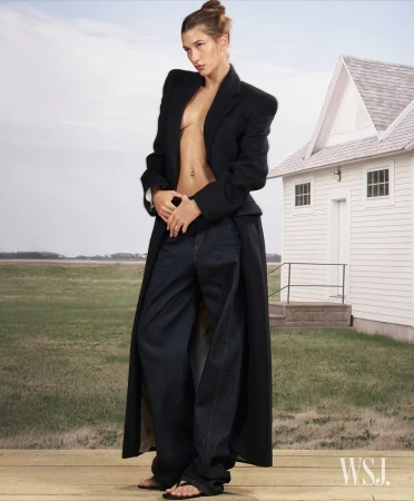 A tall, slim female model poses outdoors on a wooden deck wearing a long black coat, dark wash baggy jeans, and black sandals.