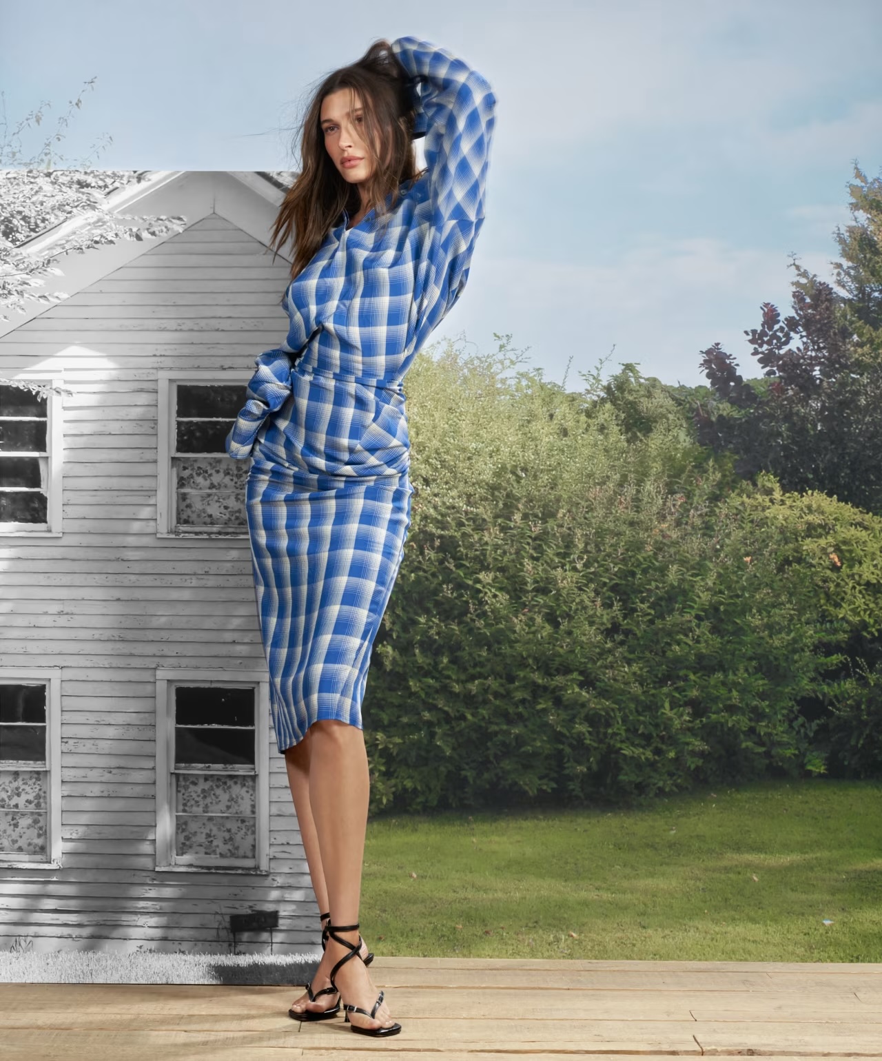 Woman poses in blue plaid skirt and top, standing on wooden floor, set against a backdrop of a house and greenery.