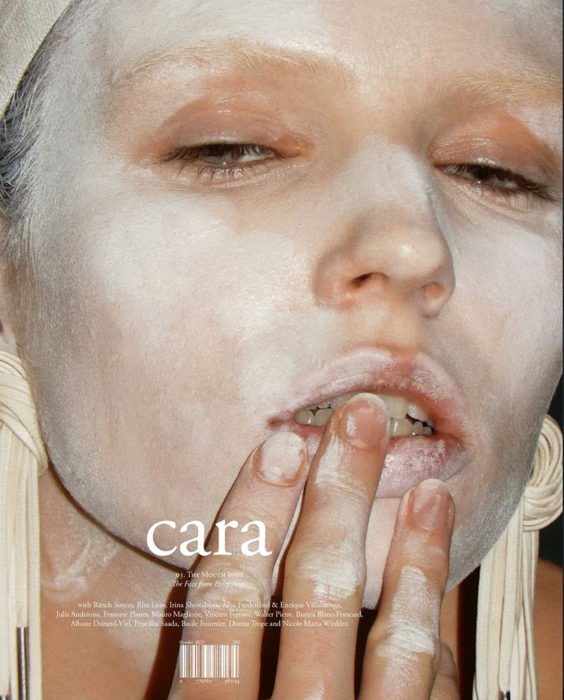 Close-up fashion shot of a model with painted white face and fingers near lips, in a studio setting with dark background, showcasing beauty and style.