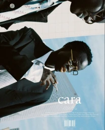 Man in a black suit, gold eyeglasses, and necktie holding a cigarette on the cover of CARA Magazine against an urban skyline.