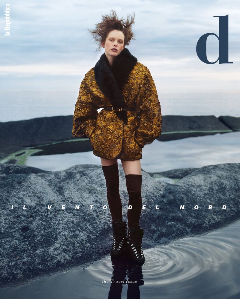 Magazine cover shot of a woman wearing a fur-collared coat, knee-high boots, standing on rocks in a pool of water with a cloudy sky.