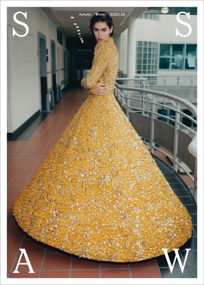 Fashion editorial shot of a woman in a gold gown, standing in an institutional hallway with large windows, for Autumn/Winter 2025-26.