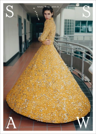 Fashion editorial shot of a woman in a gold gown, standing in an institutional hallway with large windows, for Autumn/Winter 2025-26.