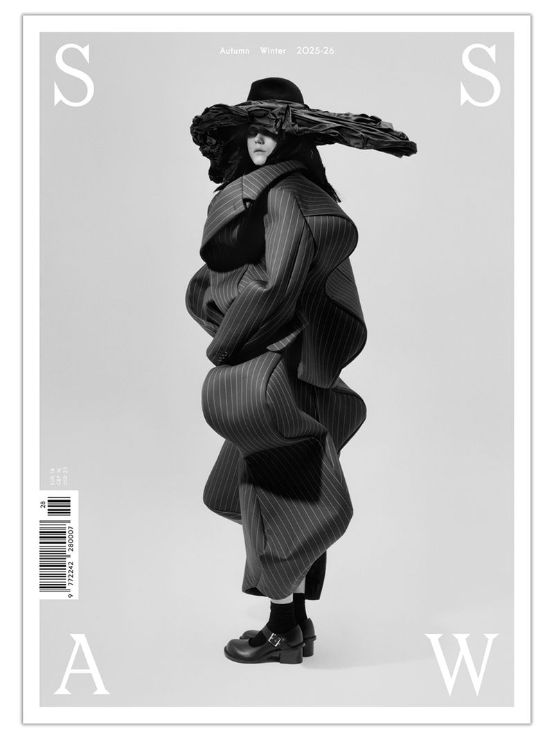 Monochrome fashion editorial shot of a model in a striped, layered coat and hat against a studio backdrop, for a magazine cover.