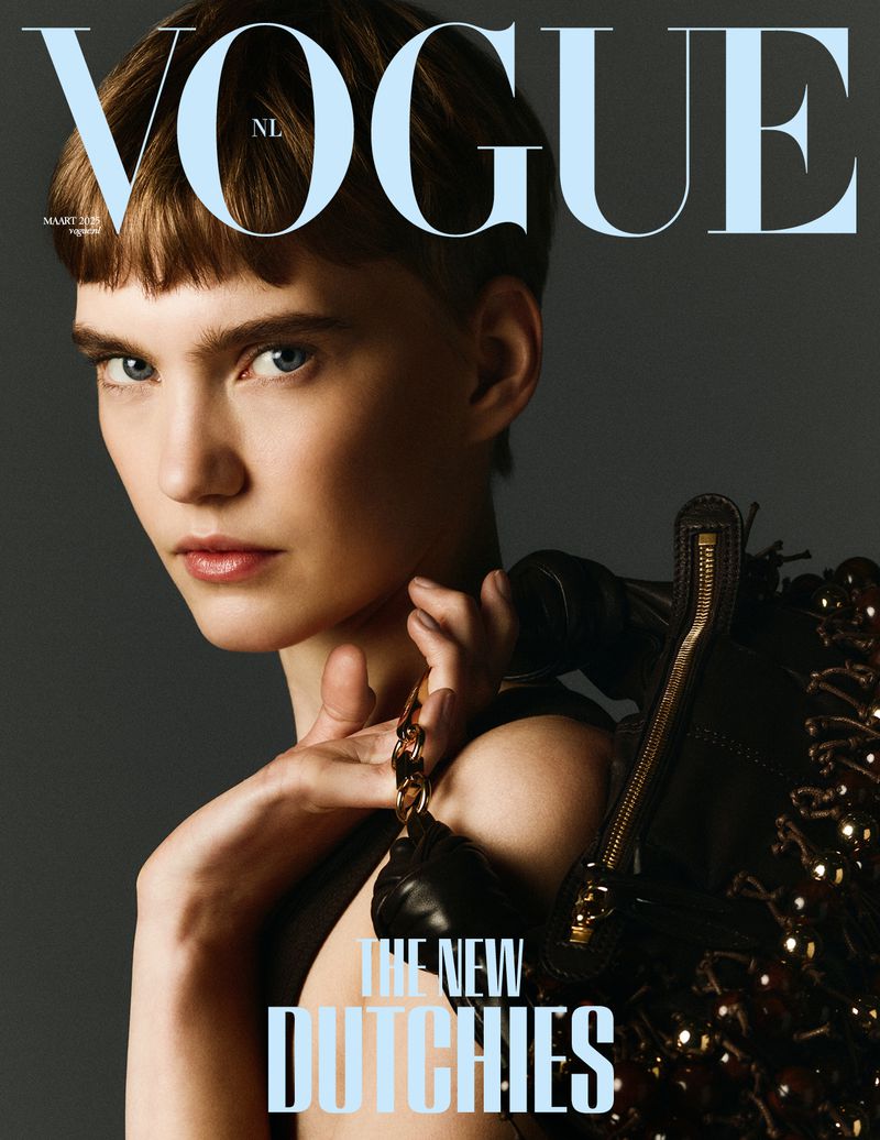 Vogue NL magazine cover featuring a woman with short hair and blue eyes, accessorized with a black handbag, set against a grey backdrop.