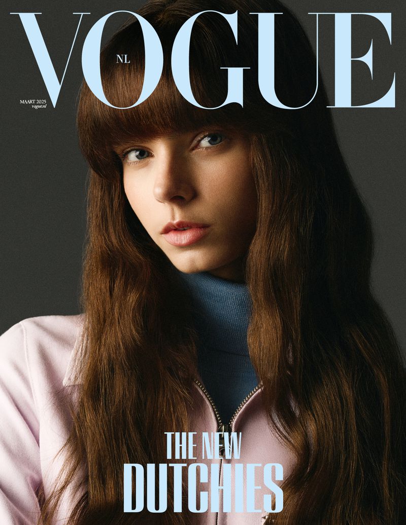 Vogue NL cover featuring a young woman with brown hair and bangs, wearing a blue turtleneck and pink jacket, against a gray background.