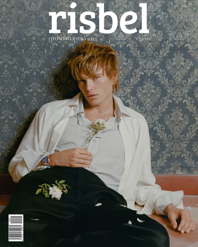 Magazine cover featuring a blond male model in an open white shirt holding a white flower, set against a patterned wall.
