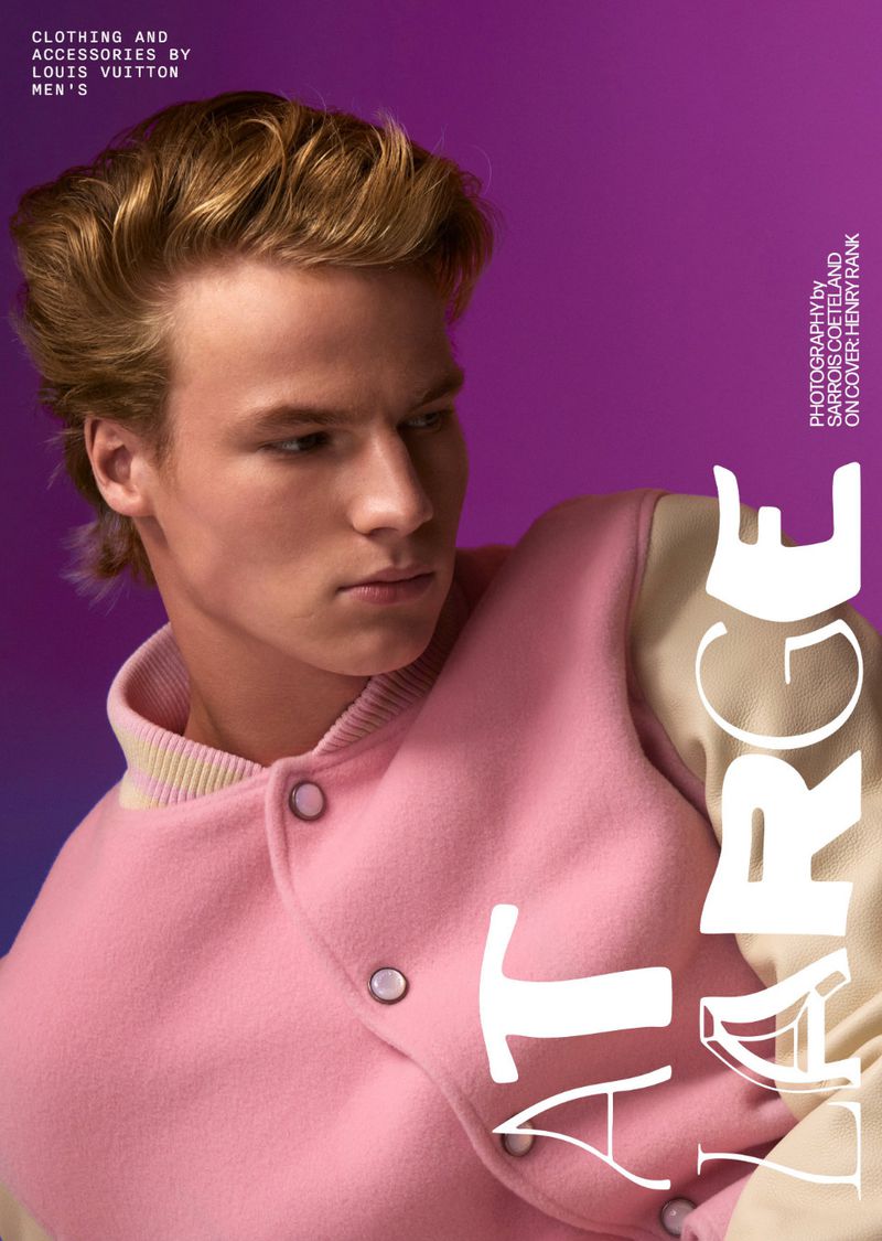 Close-up fashion shot of a young adult male with blonde hair, wearing a pink varsity jacket in front of a purple studio backdrop.