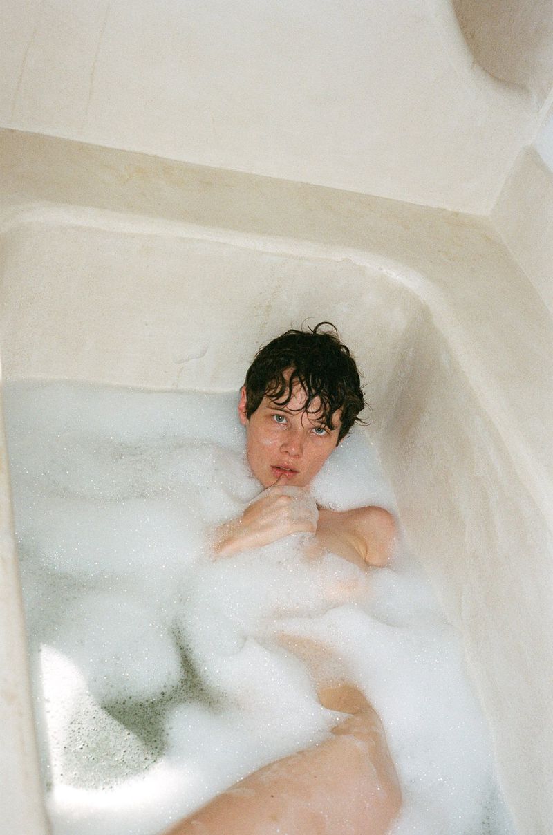 Young adult soaking in a bubble bath inside a white bathtub, looking directly at the camera with a neutral expression in a bathroom.