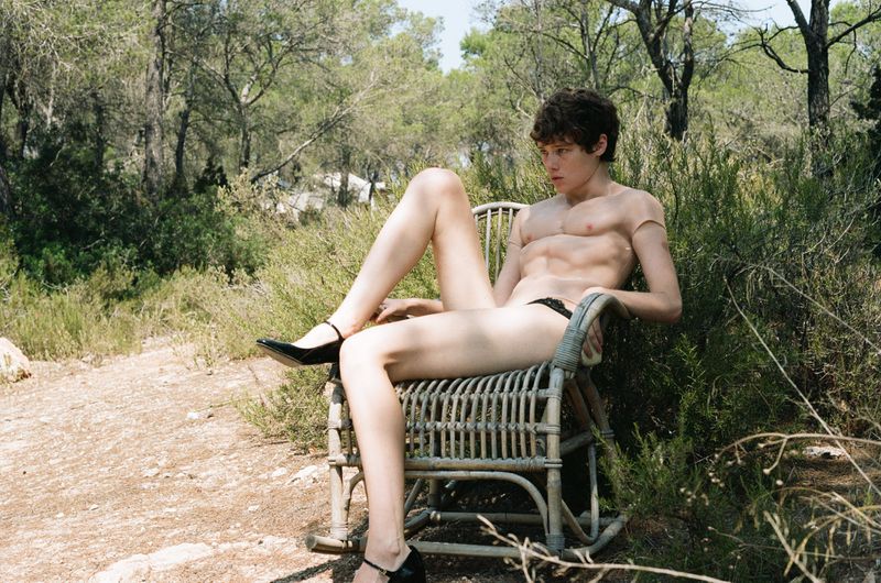 Young adult sits on woven chair in forest wearing heels and brief. Neutral expression. Ground and tree background. Outdoor setting.