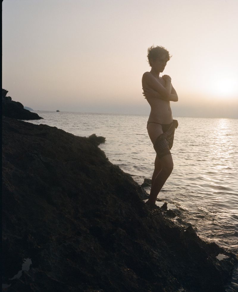 Adult female poses on a rocky beach with the sun setting over the ocean. She is facing away from the ocean and has her arms crossed.
