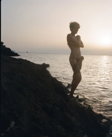 Adult female poses on a rocky beach with the sun setting over the ocean. She is facing away from the ocean and has her arms crossed.