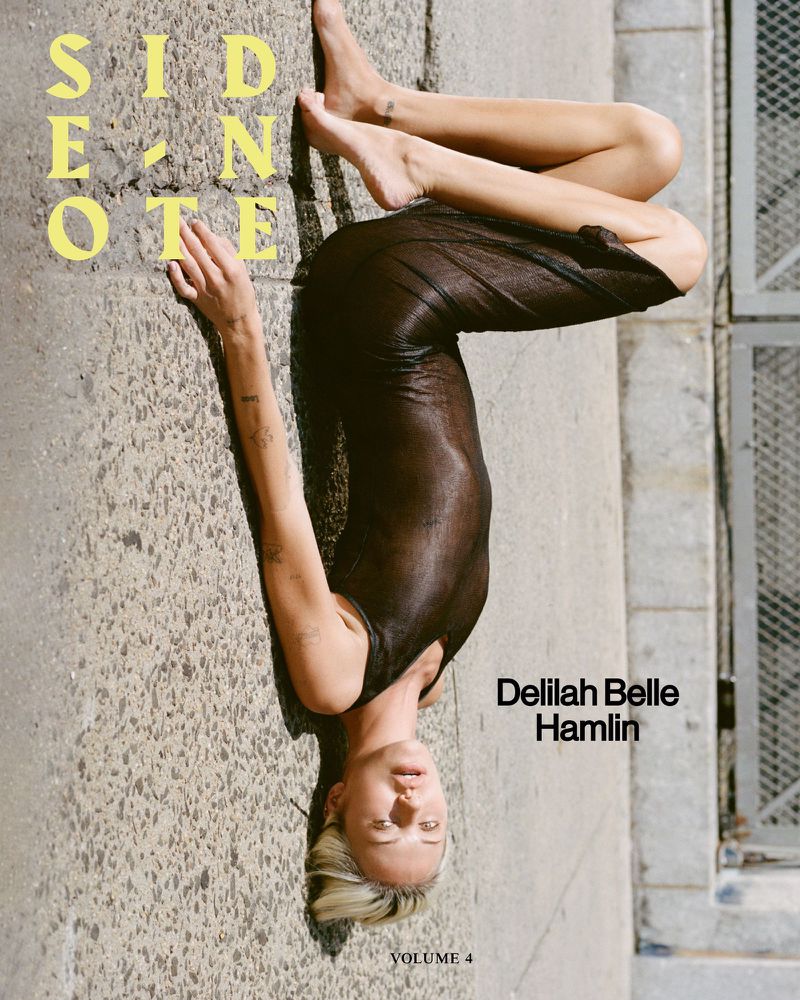 Upside-down view of Delilah Belle Hamlin on a city street, wearing a sheer dark dress against weathered concrete and building facade.