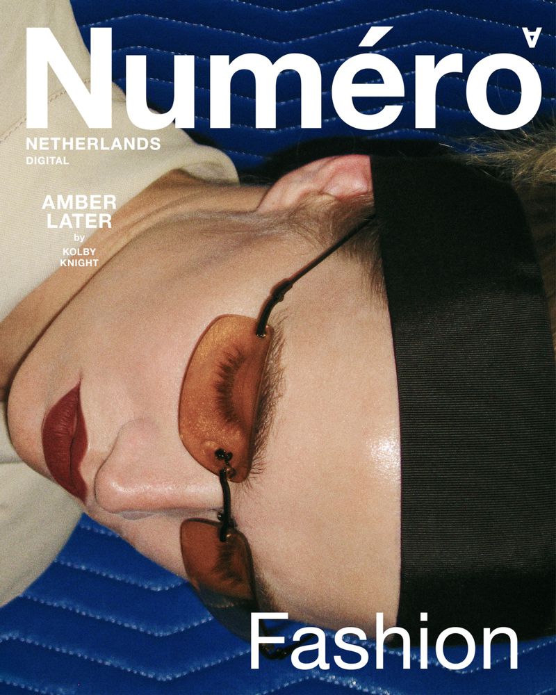 Model Amber Later wearing tinted rimless sunglasses, dark lipstick, and a black headband while resting on a royal blue textured surface.
