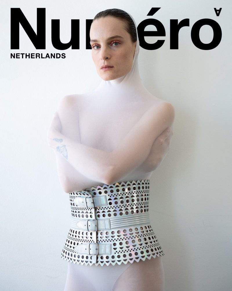 Fashion editorial featuring a slender adult woman posing in a translucent white bodysuit and a light gray, buckled, cutout corset.