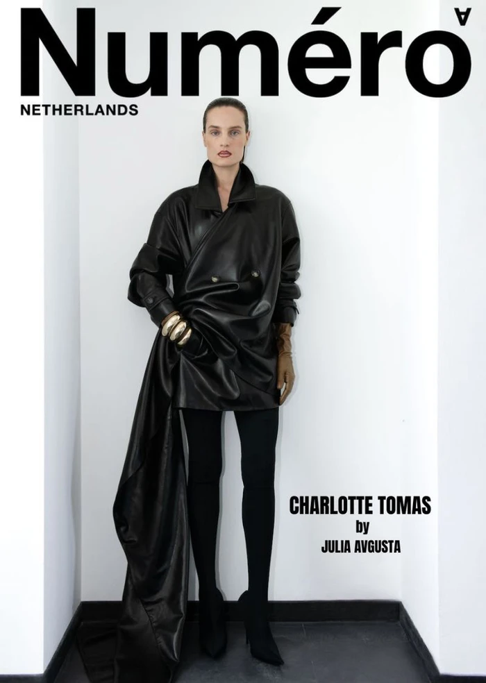 Model Charlotte Tomas poses in a black draped leather coat dress and gold bangles against a white studio corner.