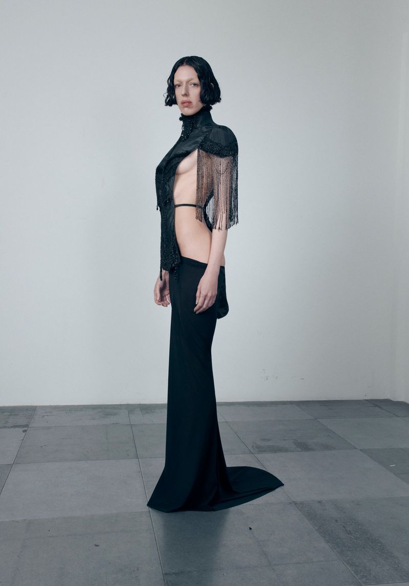 Elegant woman in black couture dress with shoulder detail poses in a bright studio with grey floor tiles, side view, fashion editorial.