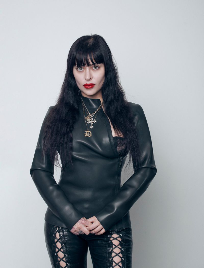 Adult female with long, dark hair, wearing a black leather jacket and pants, and standing against a plain white studio backdrop.