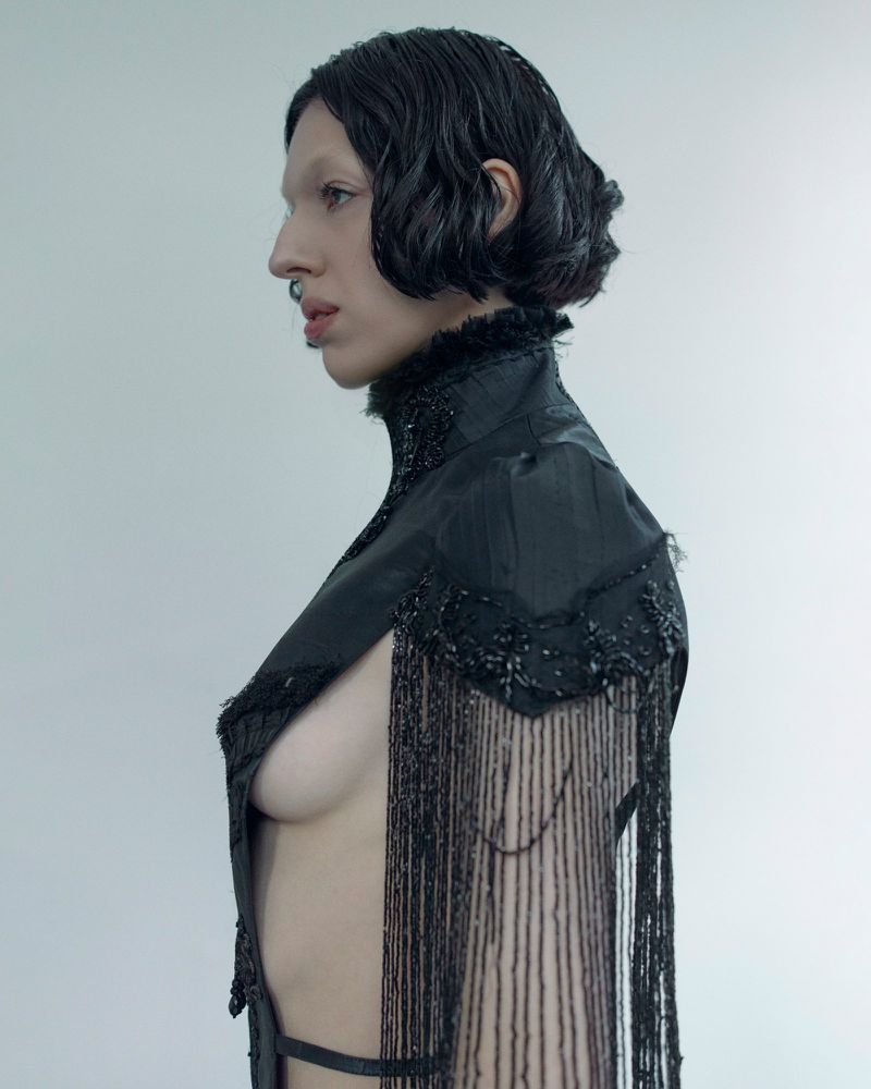 A bare-chested individual with short dark hair and a nose ring poses sideways, wearing a black fringed cape against a plain background.