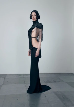 Model posing in an avant-garde Dilara Findikoglu black cutout gown with shoulder fringe against a plain wall on a tiled floor.