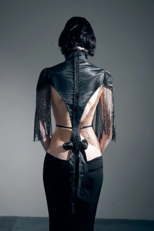 Back view of a model wearing an avant-garde backless black top with fringe sleeves and a long skirt against a grey backdrop.