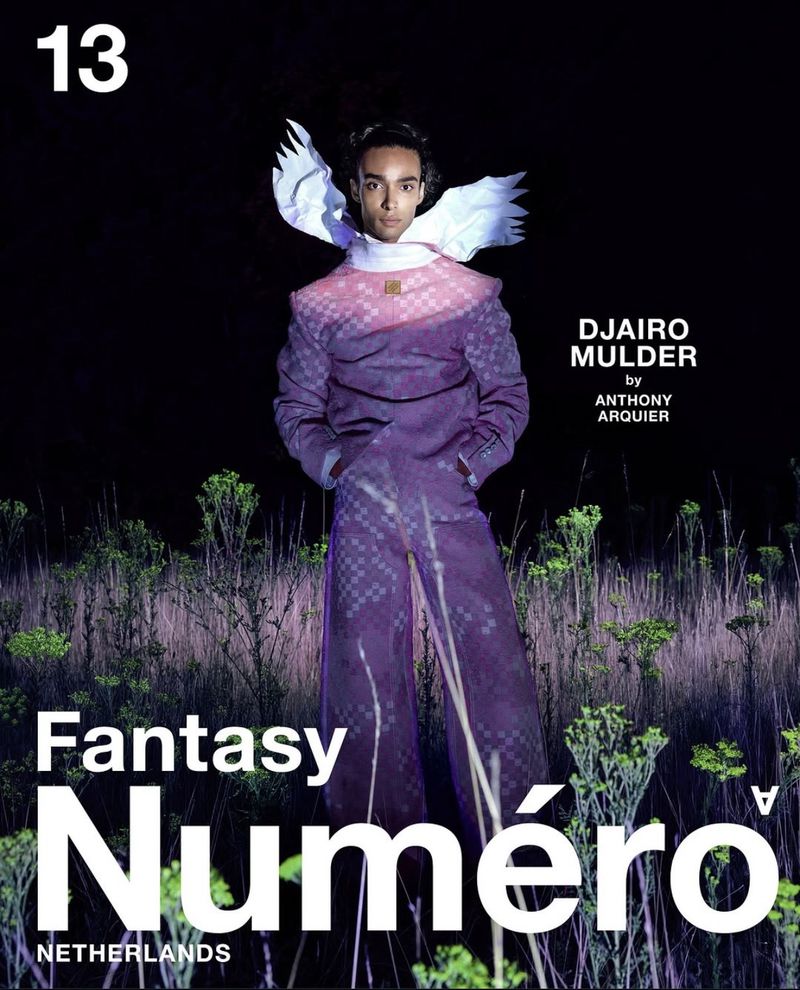Fashion editorial shot. A model wearing a purple suit with white wings stands in a field of weeds for a magazine cover.