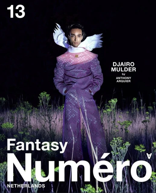 Fashion editorial shot. A model wearing a purple suit with white wings stands in a field of weeds for a magazine cover.