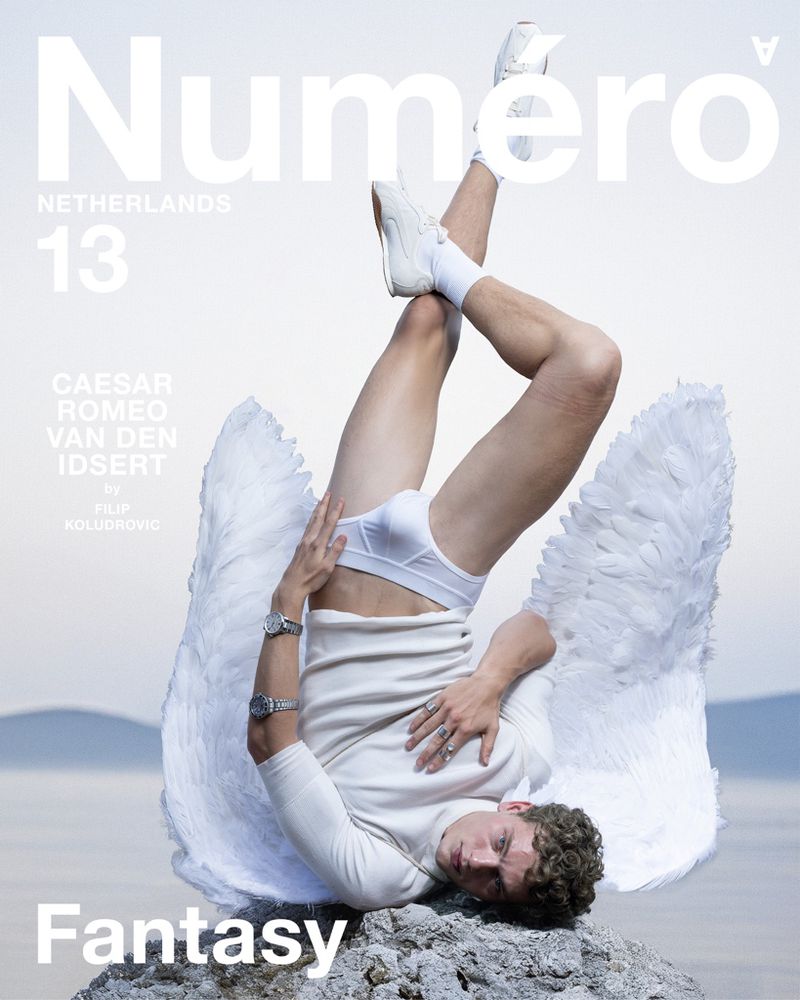 Magazine cover featuring a male model in white underwear, angel wings, and sneakers, inverting on rocks by the water with magazine text.