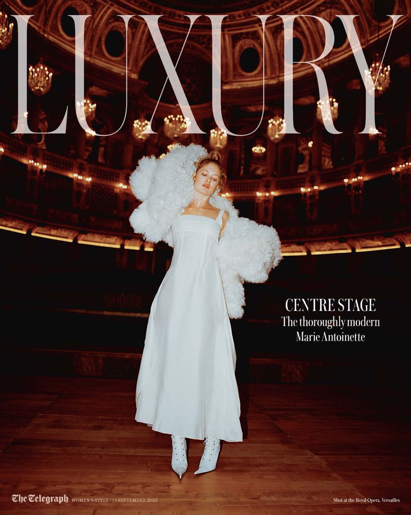 Fashion editorial shot of a woman in white gown and feather coat standing on a stage in an opera house with chandeliers.