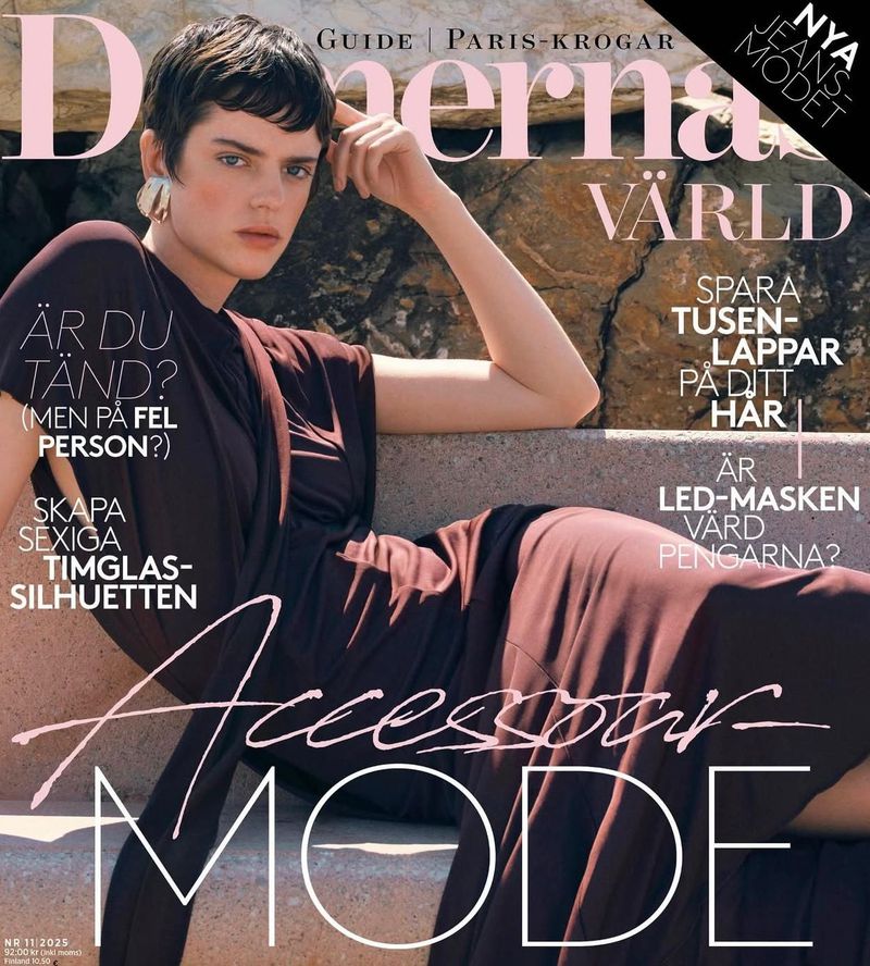 Magazine cover featuring a model with short hair wearing a burgundy dress, posing against a stone wall backdrop. Fashion editorial.