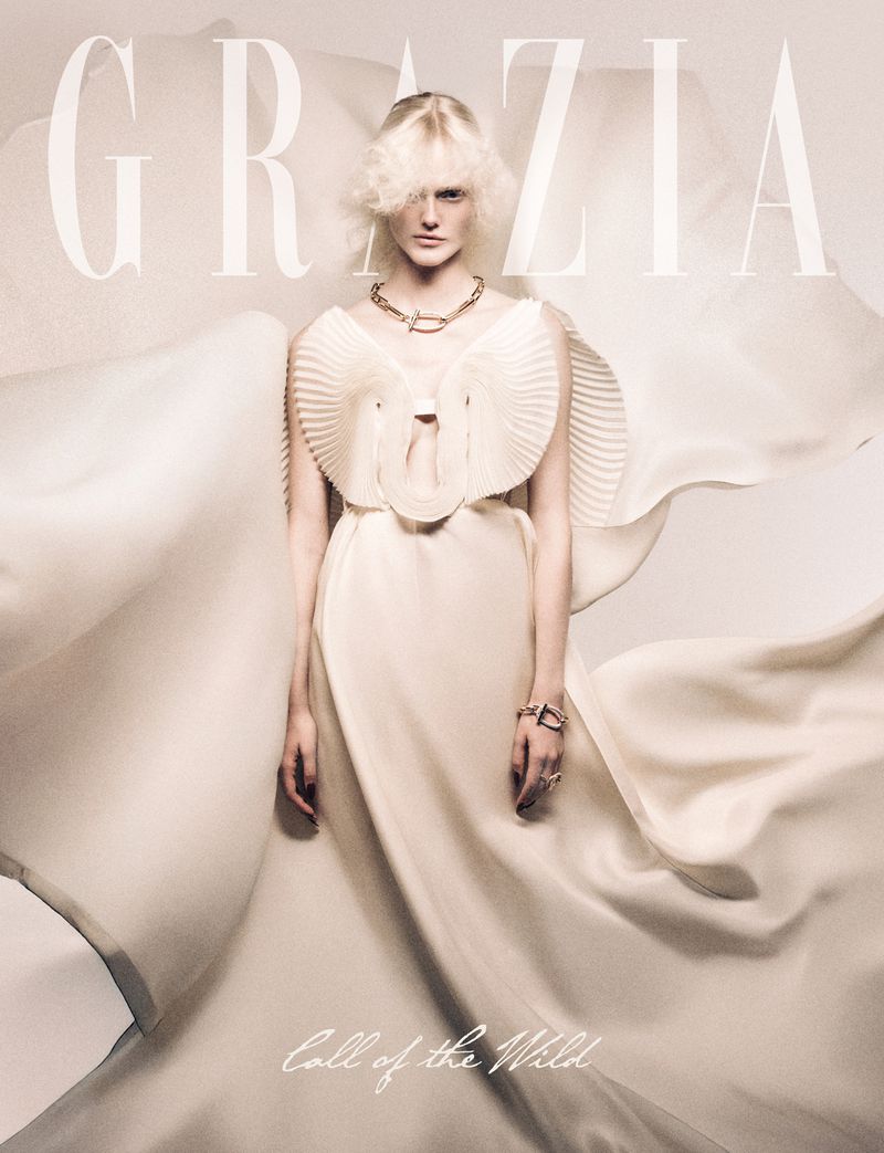 Fashion editorial shot of a stylish young woman in a white dress with a frilled collar, accessorized with jewelry and designer magazine logo.