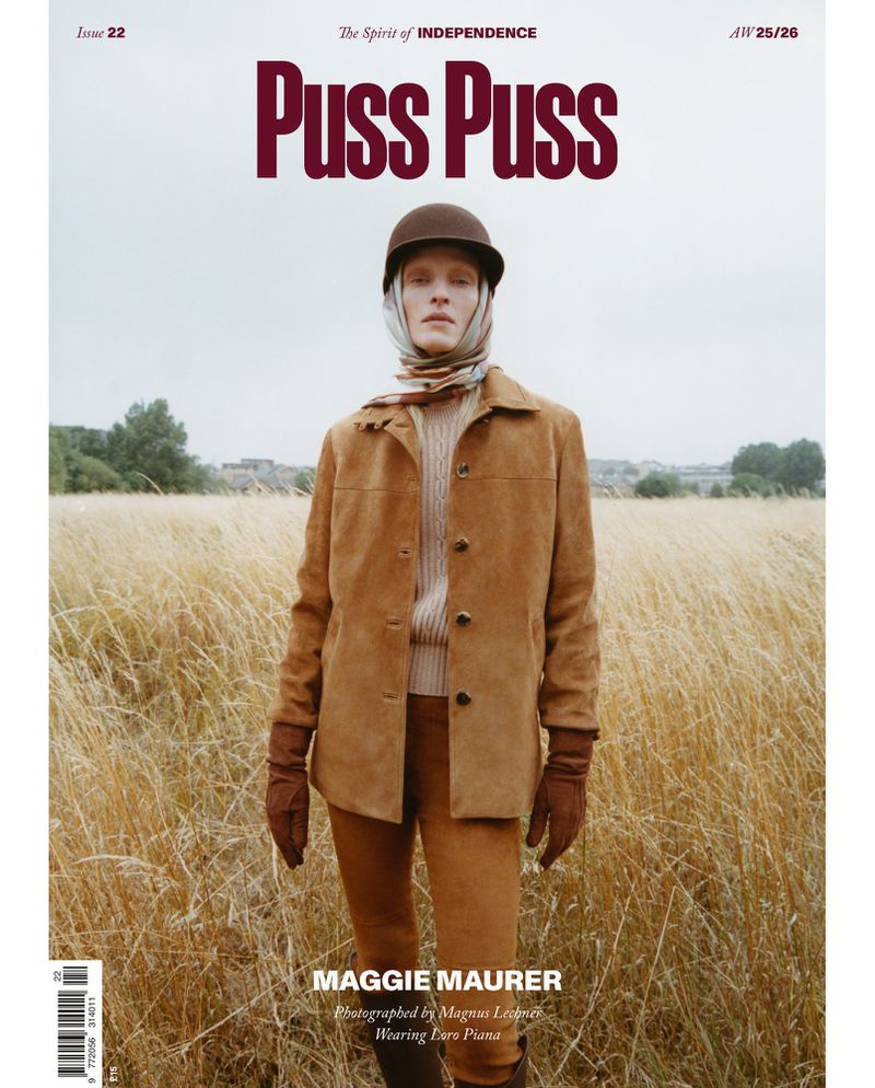 Magazine cover featuring a full-length portrait of a model in a brown jacket, helmet, and scarf, standing in a field of tall grass.