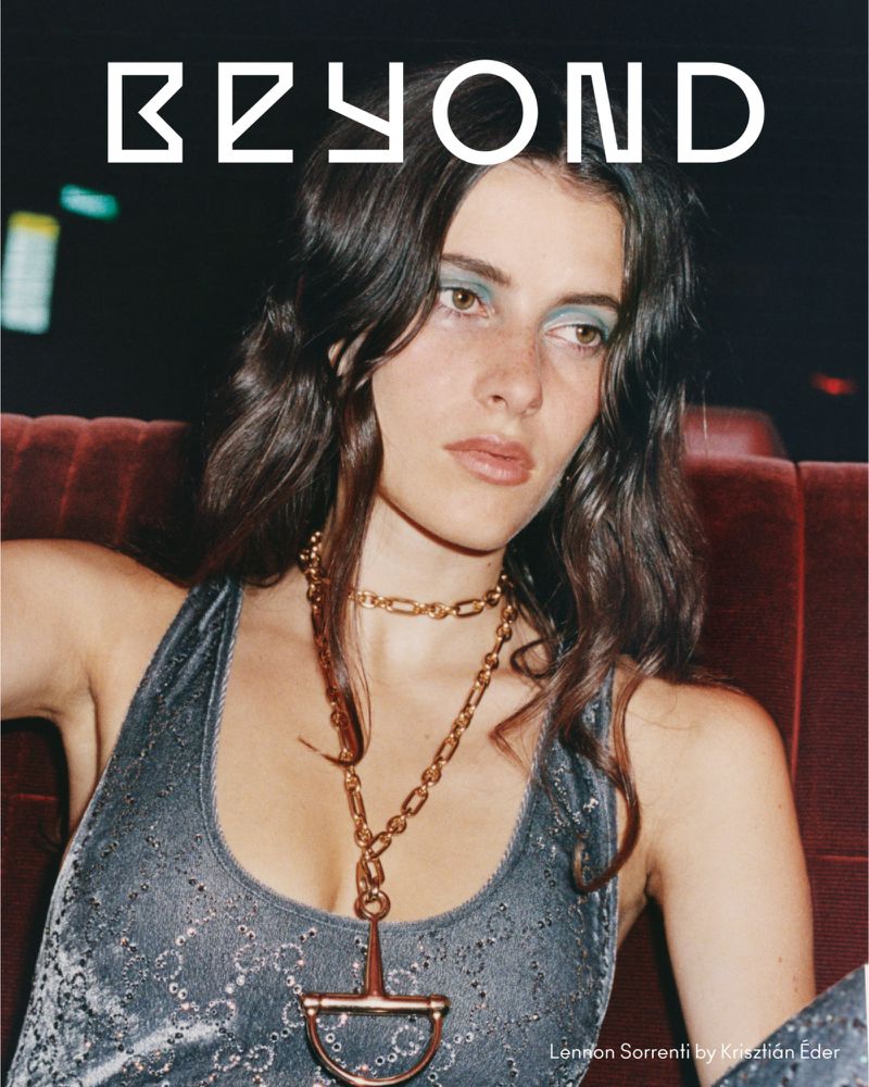 Editorial fashion shot of a brunette woman wearing a metallic top and gold jewelry, posing in a dimly lit interior space.