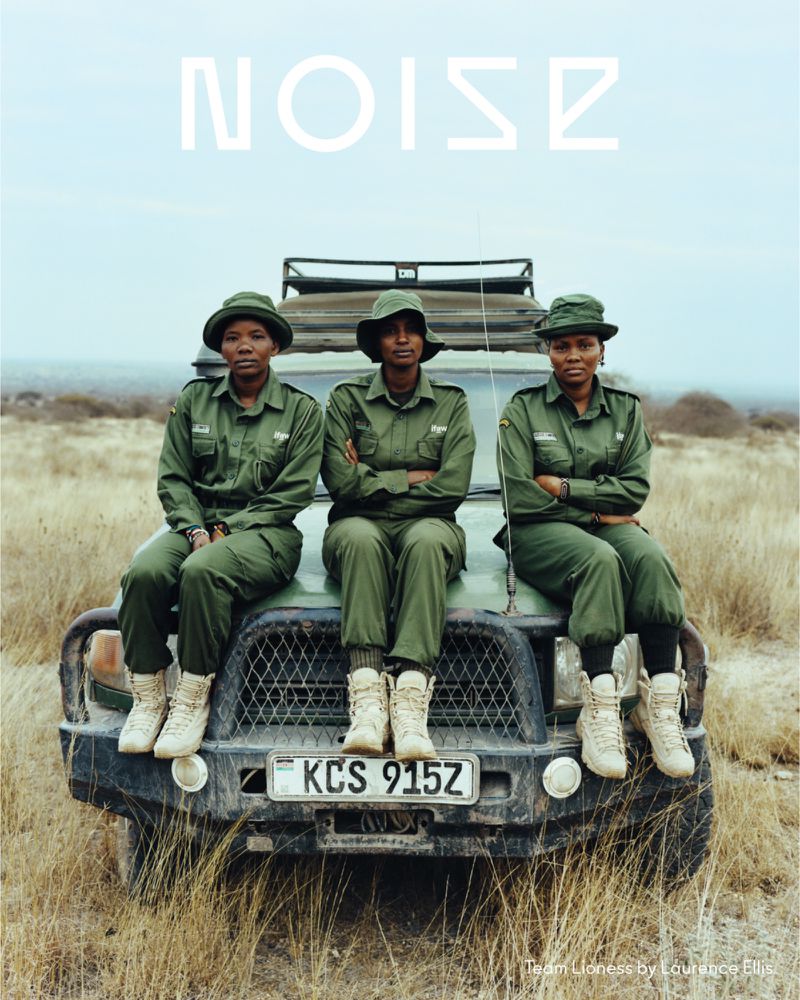 Three women in green uniforms and hats are sitting on the hood of a car. They are in a grassy, outdoor area with a clear, light blue sky.