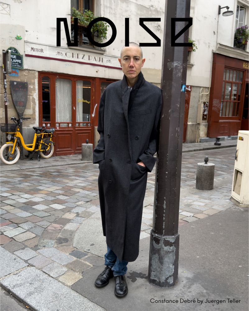 Woman in long black coat standing by a pole on a cobblestone street with building and yellow bicycle in the background in Paris.