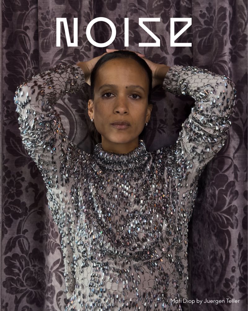 Elegant portrait of a woman with hands behind her head, wearing a beaded top, against a decorative fabric backdrop. Fashion portrait.
